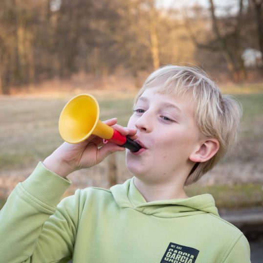 A child trumpets into a colourful plastic fanfare in front of a forest and meadow. Friedenstab Kunststofftechnik's own products are fun, as this picture shows.